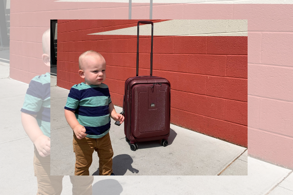 A toddler boy is standing by a rolling suitcase