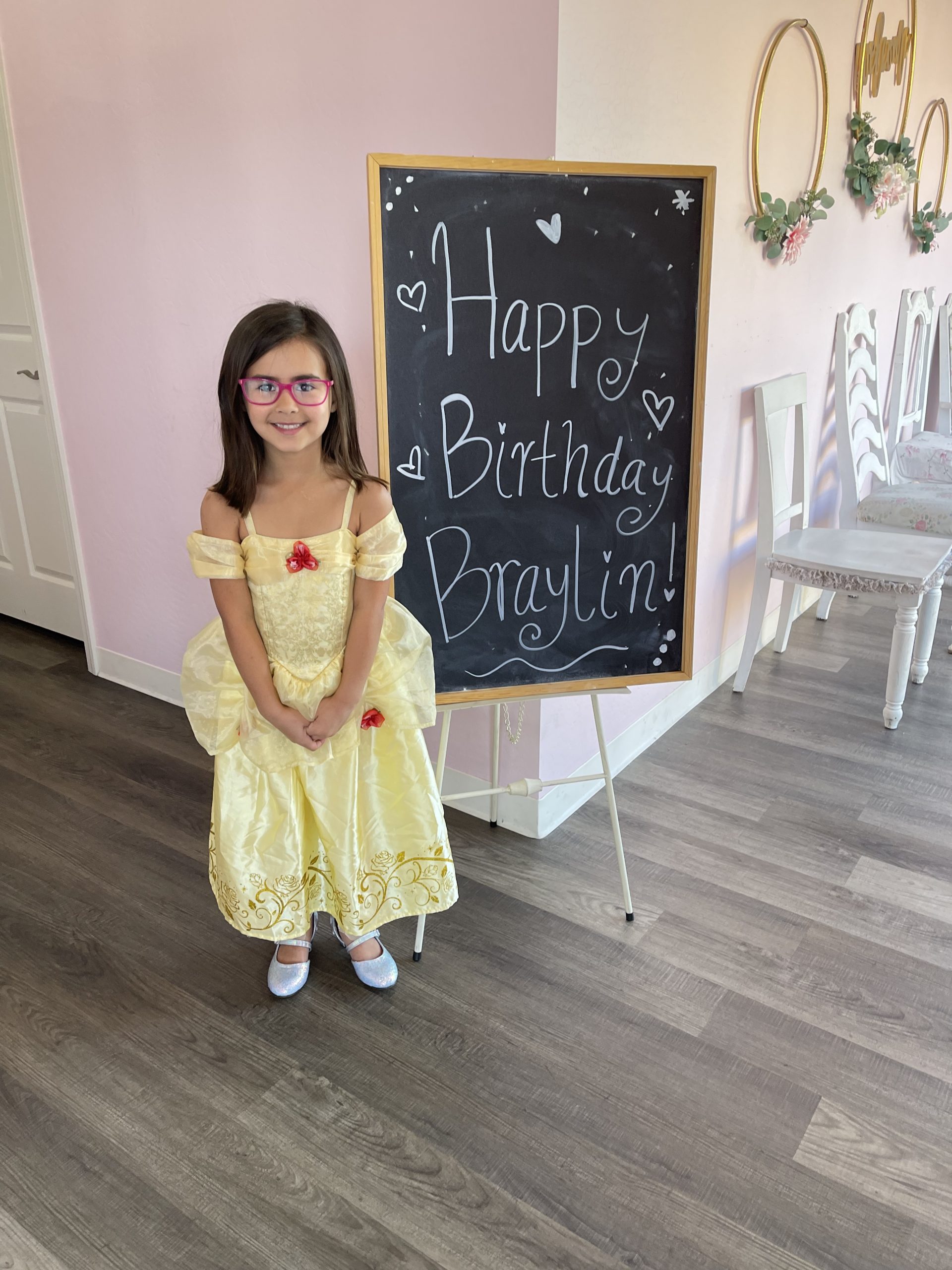 Little girl dressed up as Belle, standing next to a chalkboard sign that reads "Happy Birthday Braylin"