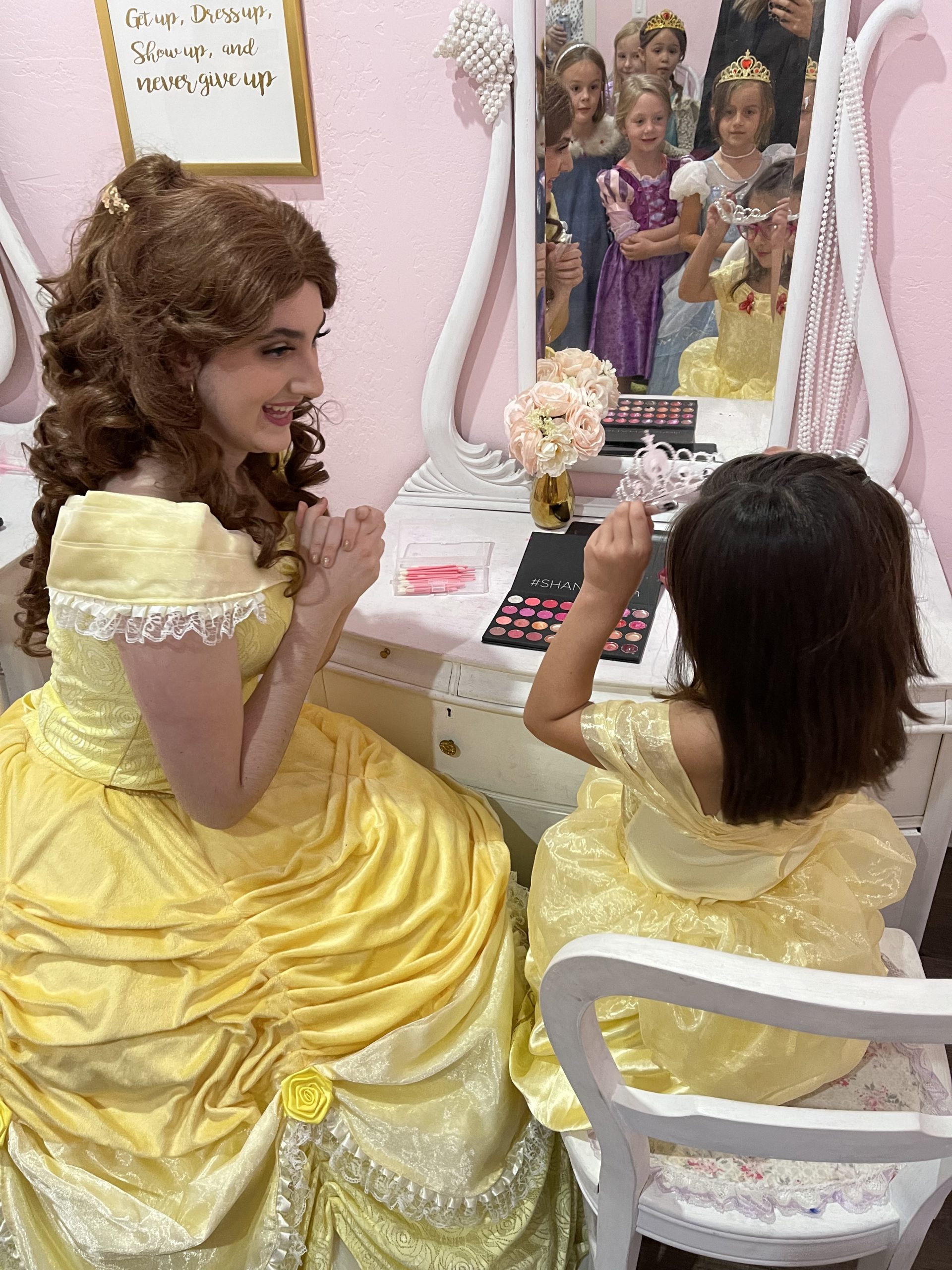 Belle and little girl dressed as Belle sitting in front of a makeup table with the reflections of other little girls dressed a princesses in the mirror.