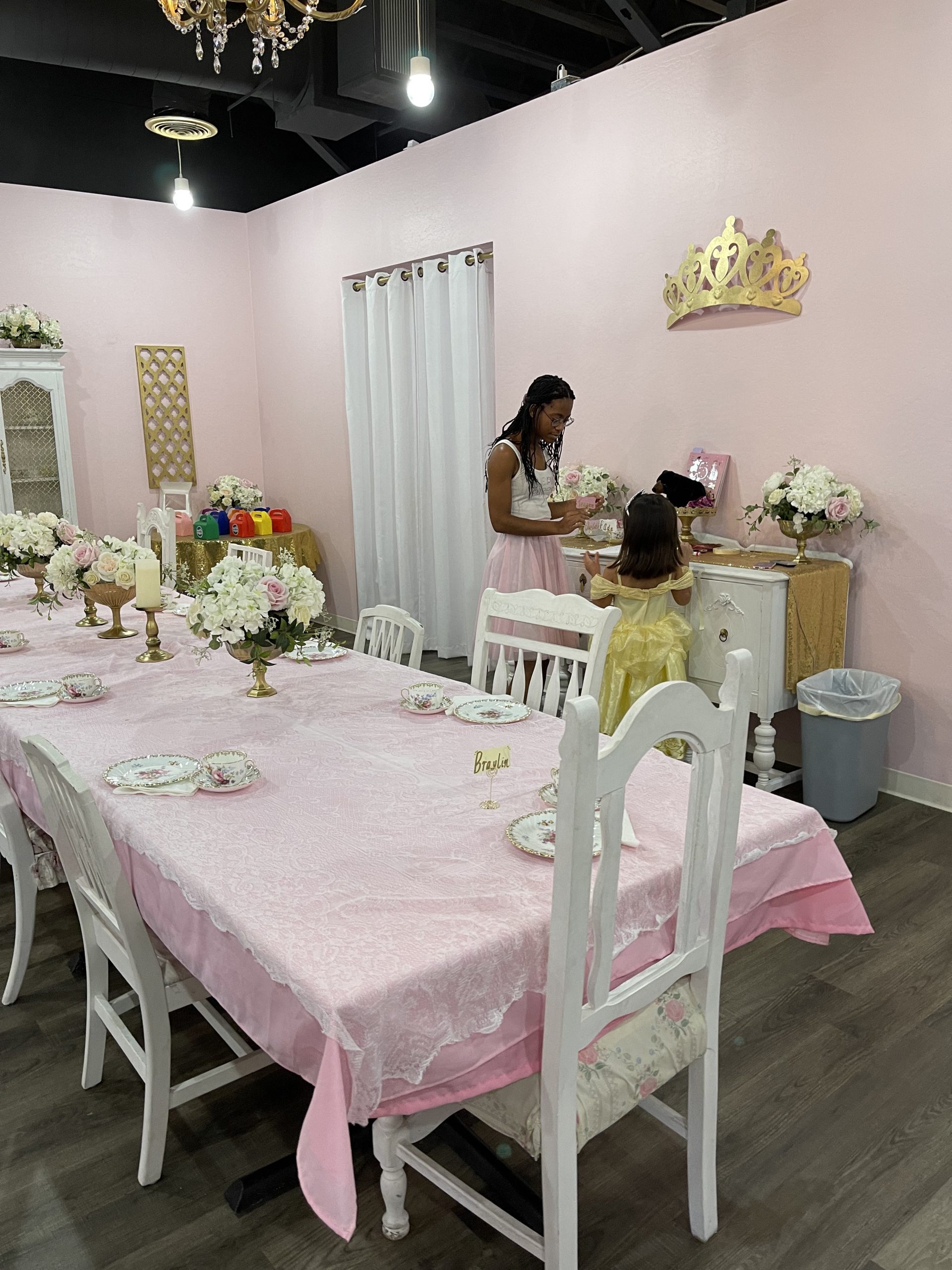 Large table set for a tea party with a little girl dressed as Belle standing next to a princess helper at a birthday party.