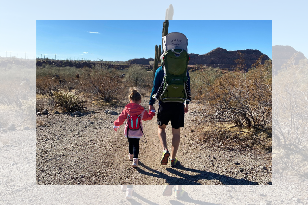 A family hiking in the Arizona desert. Dad holding the hand of a little girl while carrying a backpack with another child inside.